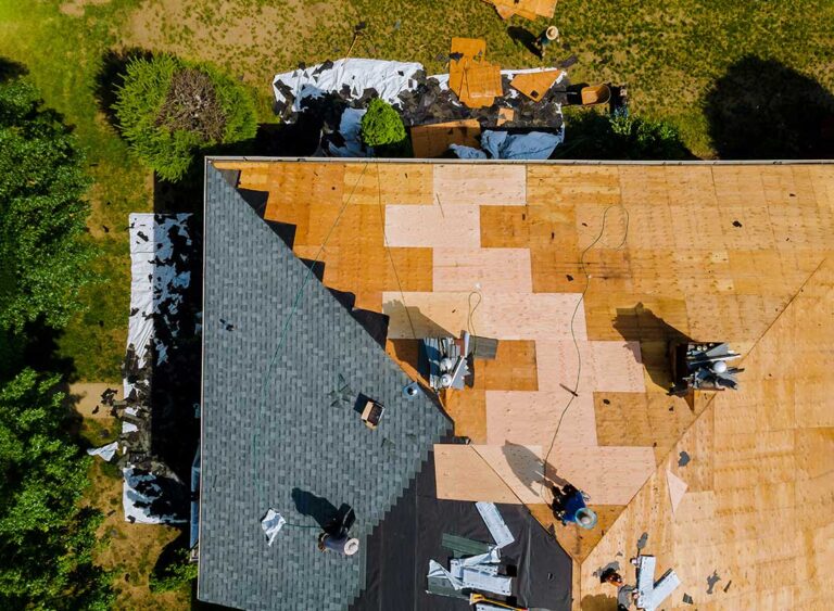 A worker applies a white protective coating to a low-slope roof beside roofing tools and drainage equipment.
